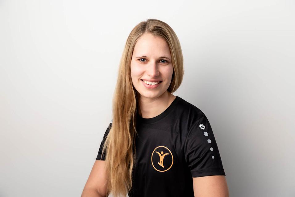 L&auml;chelnde Frau mit langen blonden Haaren tr&auml;gt ein schwarzes T-Shirt mit Logo und sitzt vor einem hellen Hintergrund.