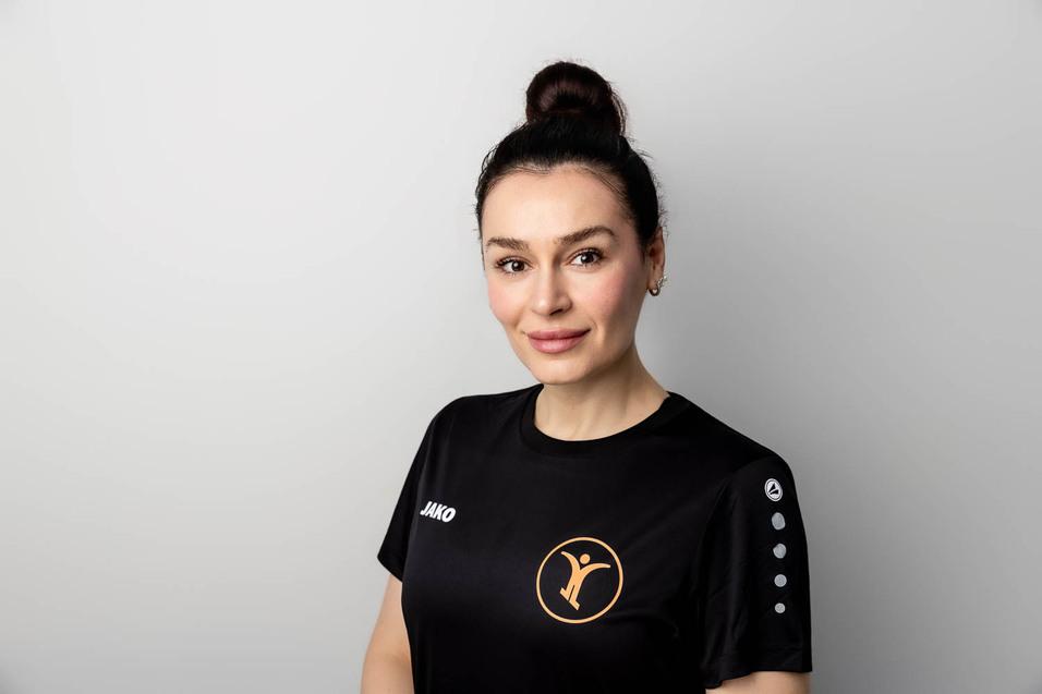 Portr&auml;t einer Frau in schwarzem Sportshirt mit Logo, l&auml;chelnd vor hellem Hintergrund.