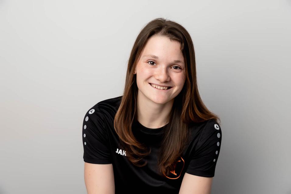 Junge Frau mit langen Haaren l&auml;chelt in schwarzem Sportshirt vor einem neutralen Hintergrund.