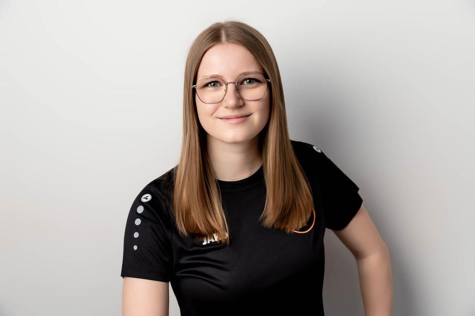 Eine junge Frau mit langen, blonden Haaren und Brille l&auml;chelt in einem schwarzen sportlichen T-Shirt vor grauem Hintergrund.