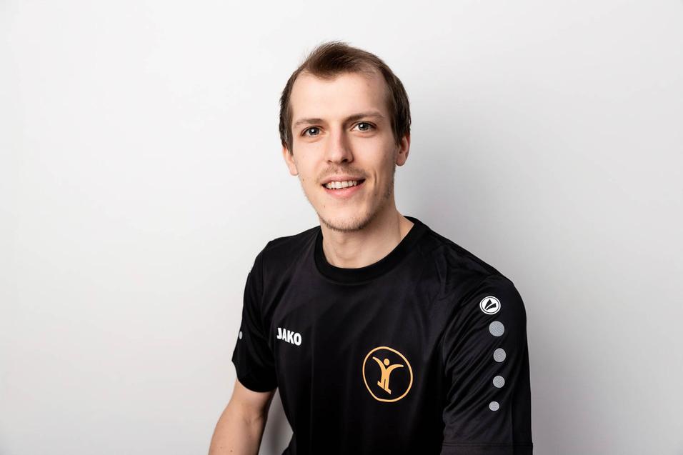 Portr&auml;t eines jungen Mannes in einem schwarzen T-Shirt mit Logo auf hellem Hintergrund, l&auml;chelnd und in seitlicher Pose.