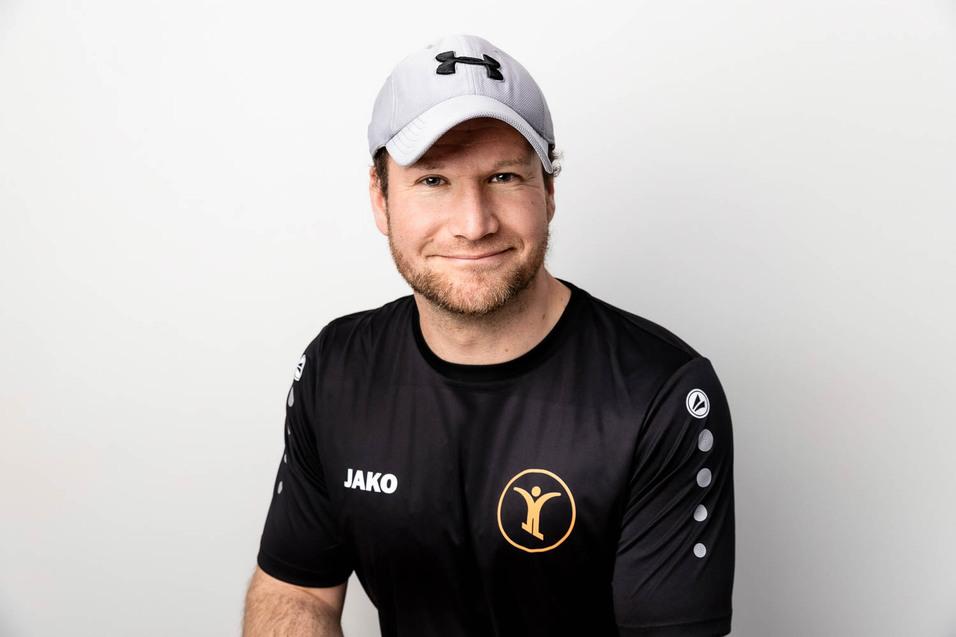 Mann in schwarzem Sportshirt mit Logo und grau-wei&szlig;em Cap l&auml;chelt vor wei&szlig;em Hintergrund.