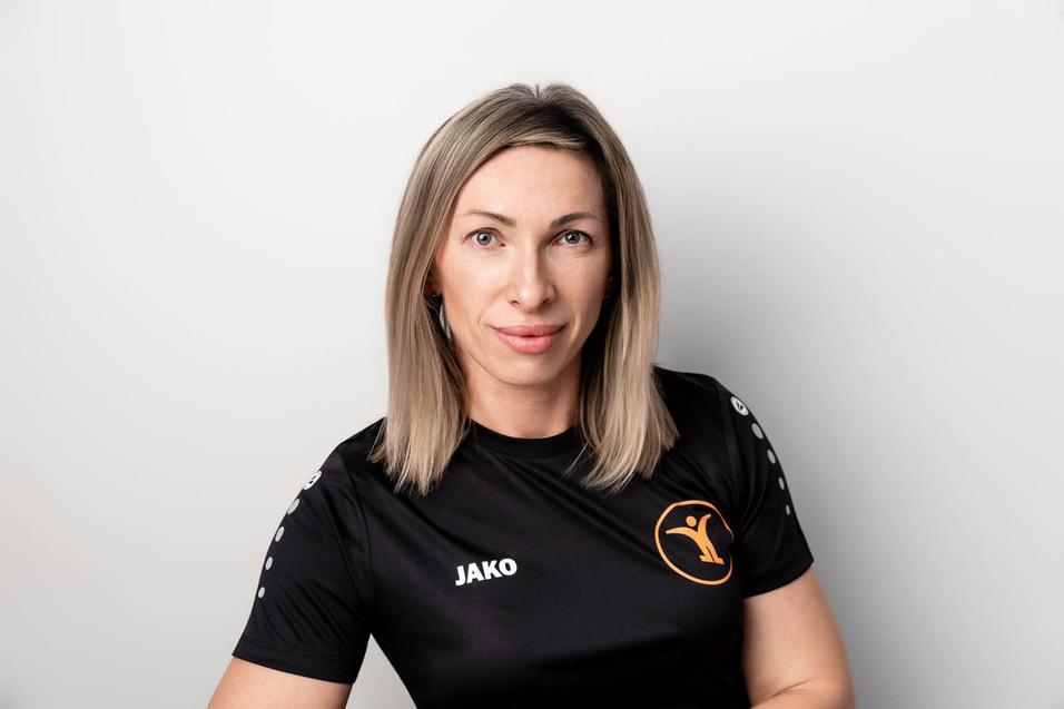 Portr&auml;t einer sportlichen Frau mit blonden Haaren, die ein schwarzes T-Shirt mit Logo tr&auml;gt, vor einem hellen Hintergrund.