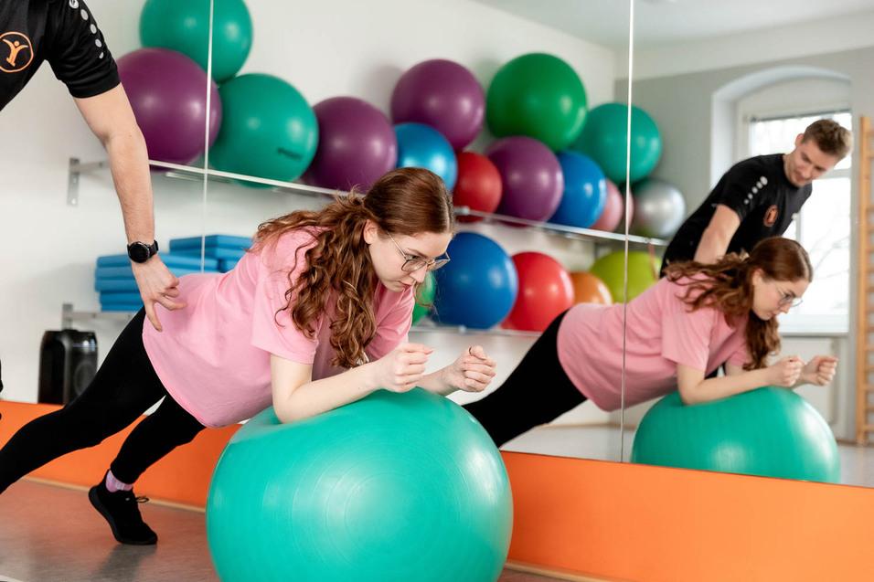 Frau mit Brille und pinkem Shirt &uuml;bt mit einer gro&szlig;en gr&uuml;nen Trainingsball, begleitet von Trainer in Sportkleidung.