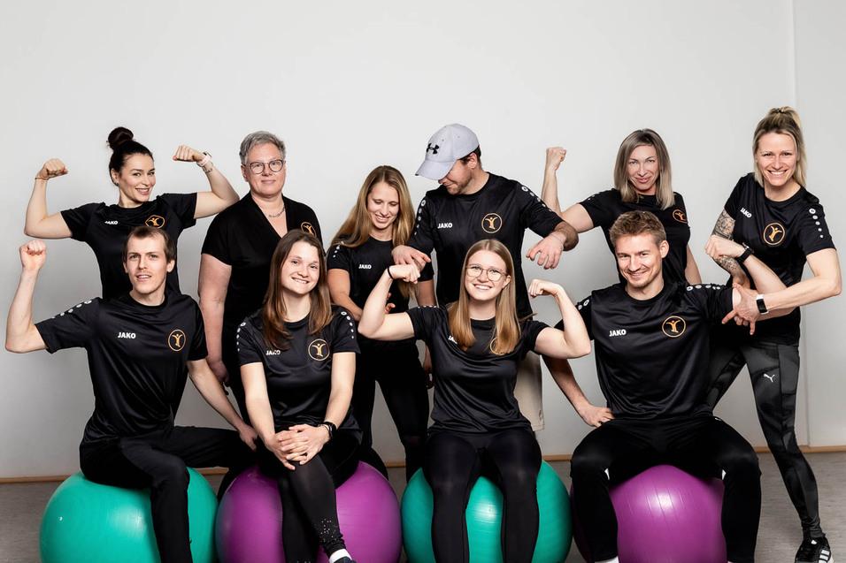 Gruppe von neun Personen in Sportkleidung, die auf Fitnessb&auml;llen sitzen oder stehen, und ihre Muskeln zeigen.