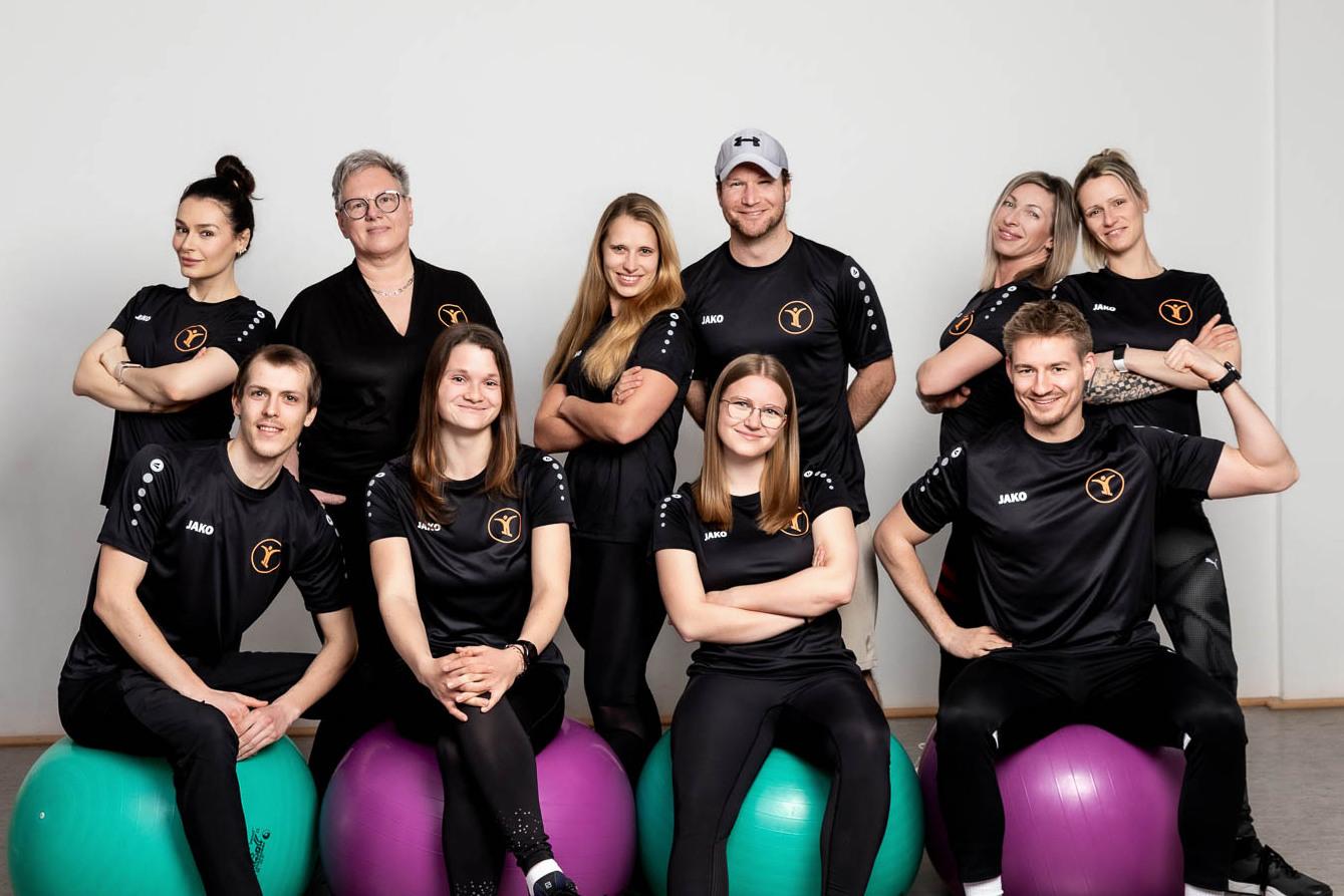 Gruppe von zehn Personen in Sportkleidung sitzt auf Fitnessb&auml;llen und posiert vor einer neutralen Wand.