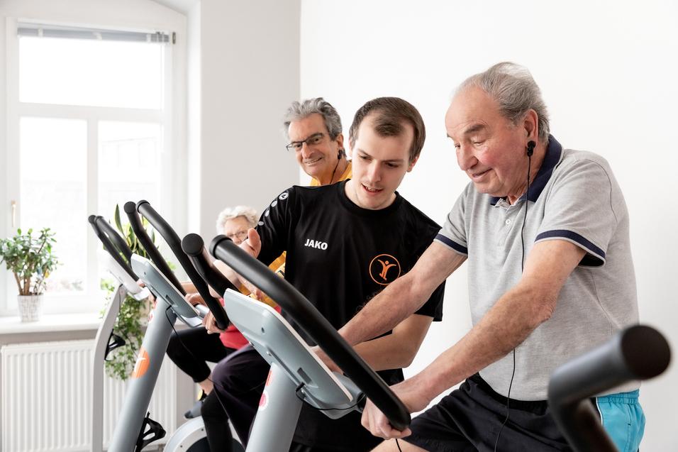&Auml;ltere Menschen trainieren an Fitnessger&auml;ten, w&auml;hrend ein junger Trainer Anweisungen gibt. Helle, freundliche Umgebung.