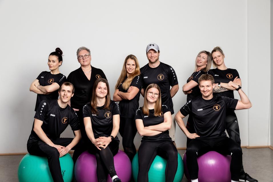 Gruppe von neun Personen in Sportkleidung, die auf großen bunten Gymnastikbällen sitzen und lächeln.
