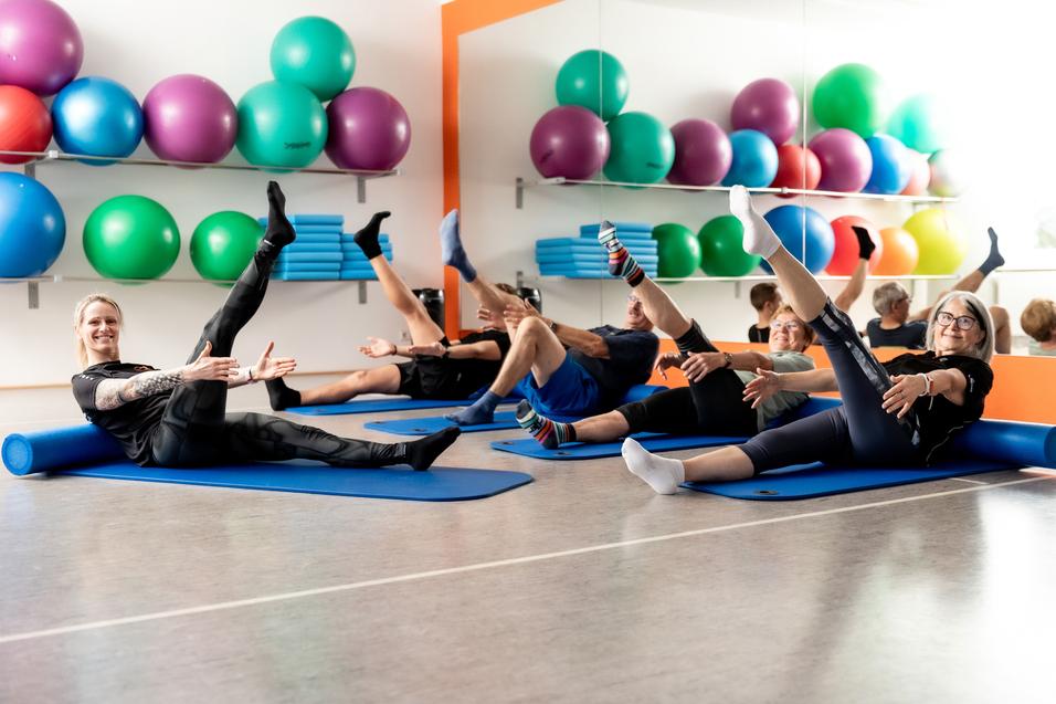 Gruppe von Fitnesssportlern f&uuml;hrt &Uuml;bungen auf blauen Matten in einem Raum mit bunten Gymnastikb&auml;llen aus.