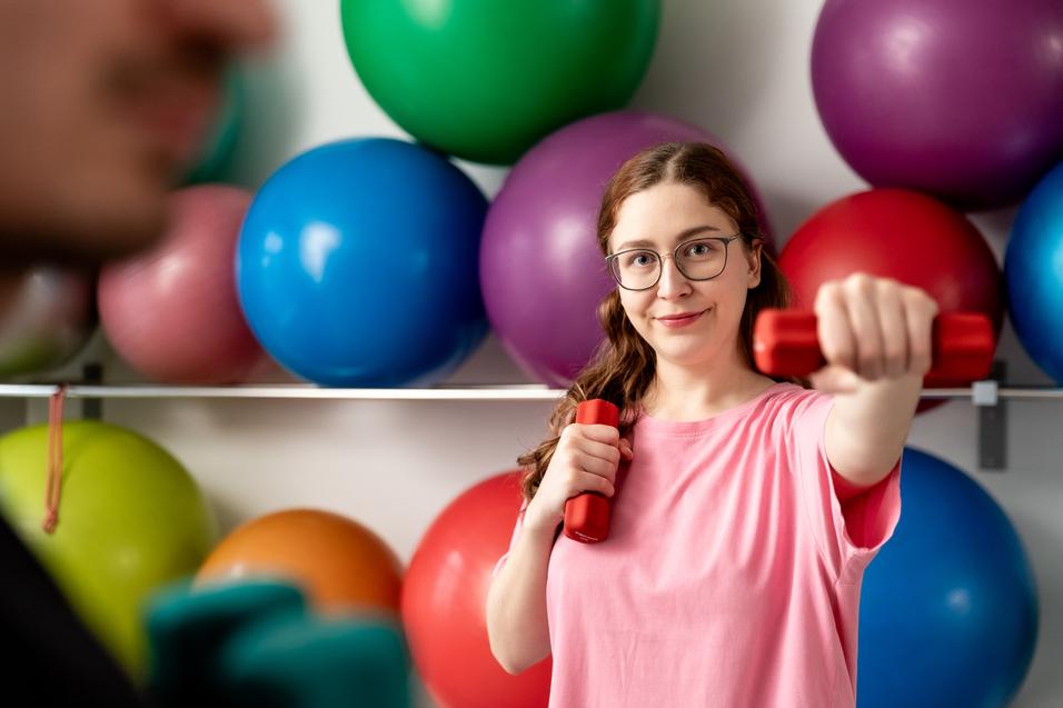 Frau mit Handgewicht in sportlicher Pose vor einer Wand mit bunten Gymnastikb&auml;llen.