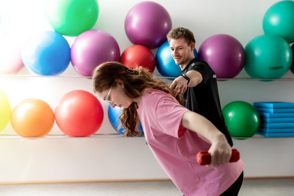 Trainer unterstützt Frau mit Hanteln beim Workout in einem Raum mit bunten Gymnastikbällen im Hintergrund.