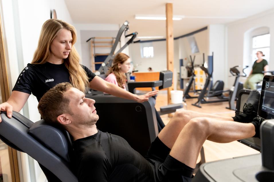 Eine Trainerin assistiert einem Mann beim Beinpressen an einem Fitnessger&auml;t in einem modernen Trainingsraum.