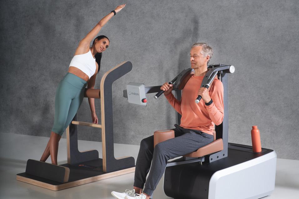 Mann im Fitnessstudio trainiert an einer Maschine, w&auml;hrend eine Frau neben ihm gedehnt. Beide nutzen moderne Ger&auml;te.