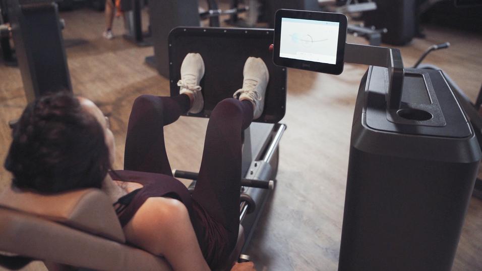 Frau trainiert an einer Beinpresse mit Display im Fitnessstudio. F&uuml;&szlig;e sind auf der Plattform, K&ouml;rper leicht zur&uuml;ckgelehnt.
