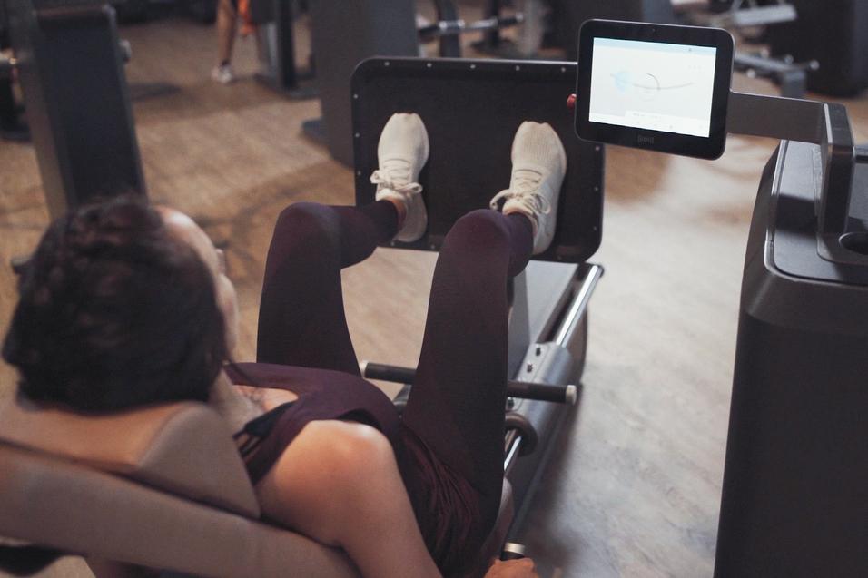 Frau trainiert an einer Beinpresse mit Display im Fitnessstudio. F&uuml;&szlig;e sind auf der Plattform, K&ouml;rper leicht zur&uuml;ckgelehnt.