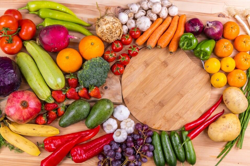 Bunte Auswahl an frischem Obst und Gem&uuml;se arrangiert um ein rundes Holzbrett auf einem Tisch.