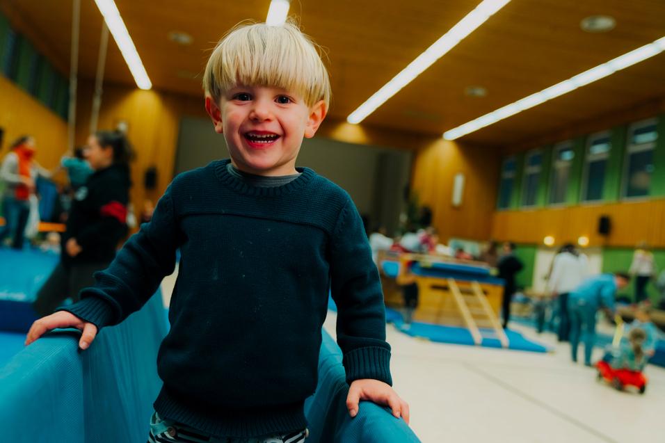 Lächelnder Junge mit blonden Haaren, der an einer blauen Turnmatte in einer Sporthalle steht, im Hintergrund sind andere Kinder aktiv.