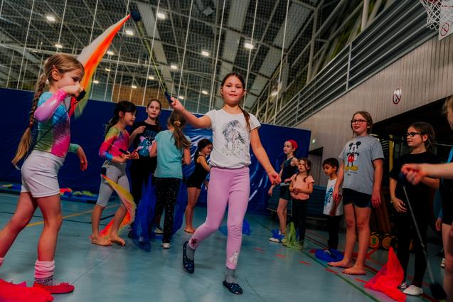 Mädchen in Sportkleidung üben mit bunten Tüchern in einer Turnhalle; Gruppenaktivität mit fröhlichen Gesichtern.