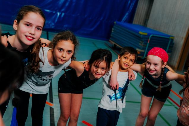 Sechs Kinder umarmen sich lächelnd in einer Sporthalle, mit Turnmatten im Hintergrund.