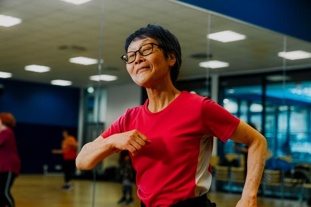 Eine lächelnde Frau in rotem Sportshirt tanzt motiviert in einem Fitnessraum vor einem Spiegel.
