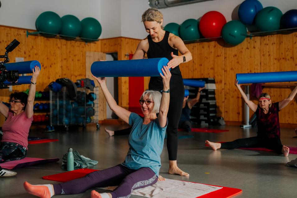 Frau hebt eine blaue Fitnessrolle über einer sitzenden Frau mit Brille in einer Gruppentrainingssession.