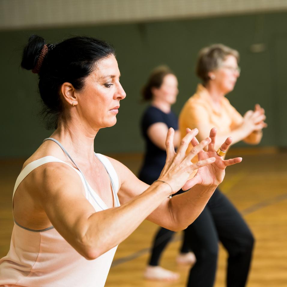 Drei Frauen führen eine Fitnessübung in einem Saal mit Holzfußboden und einer Sporttorwand im Hintergrund aus.