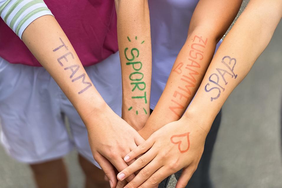 Vier Arme liegen übereinander, mit farbiger Schrift: „TEAM“, „SPORT“, „ZUSAMMEN“, „SPASS“ auf der Haut.