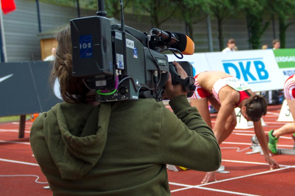 Kameramann filmt sprintende Athletinnen an der Startlinie auf einer leuchtend roten Tartanbahn.