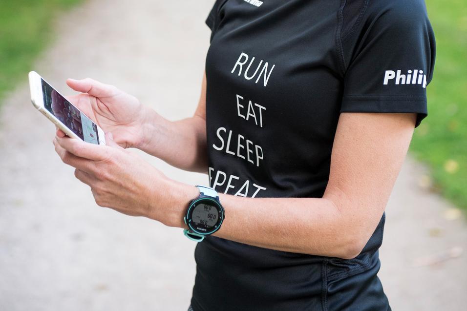 Frau mit kurzer blonder Frisur schaut auf ihr Smartphone, trägt ein Sport-T-Shirt mit Schriftzug und eine Sportuhr am Handgelenk.