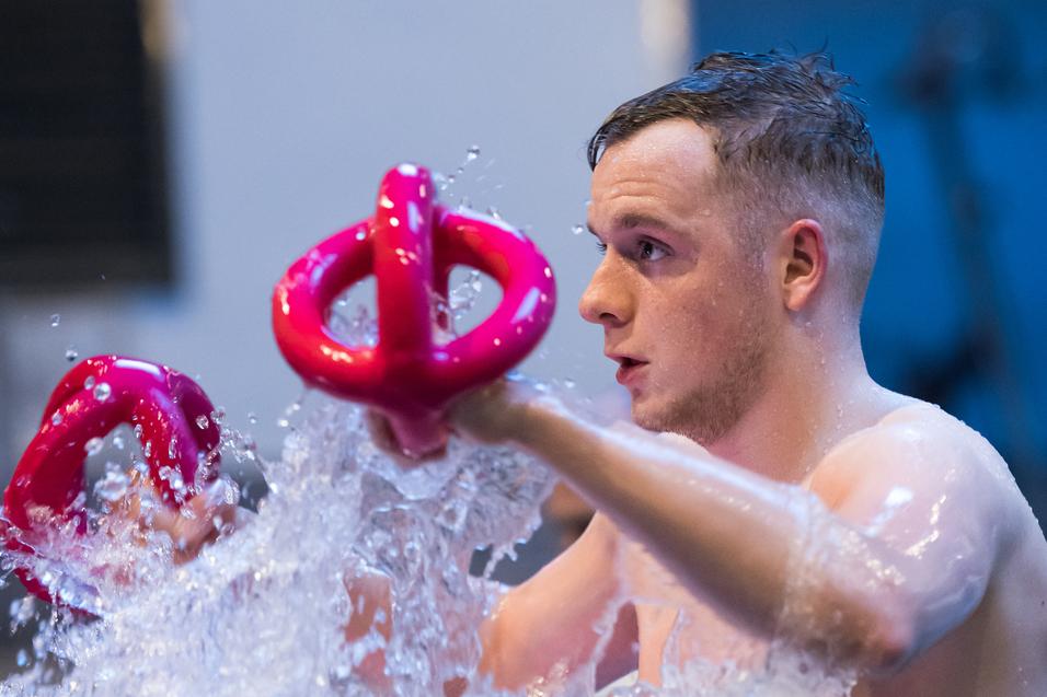 Junger Mann im Wasser hält zwei pinke Wasserhanteln und spritzt Wasser während des Trainings.