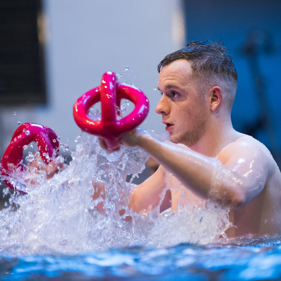 Junger Mann im Wasser hält zwei pinke Wasserhanteln und spritzt Wasser während des Trainings.